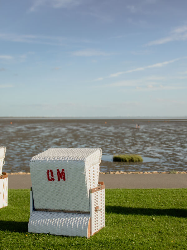 Weißer Strandkorb mit roten Initialen auf der Deichwiese, dahinter das Watt